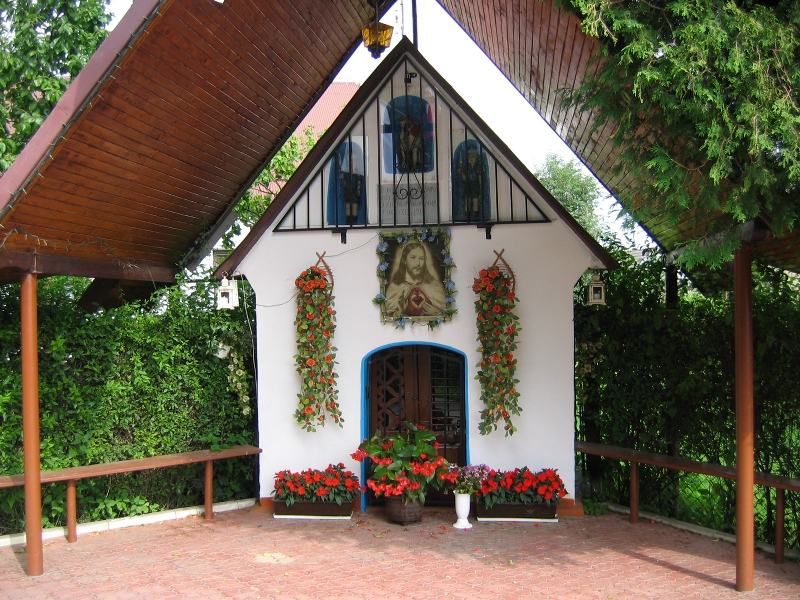 Kapliczka w Małej Wsi - niewielka kapliczka z obrazem Jezusa Chrystusa pośrodku, przystrojona kwiatami, przed kapliczką zadaszone miejsce z ławkami do siedzenia, w tle zielony żywopłot
