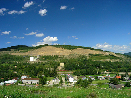 Panorama miejscowości, na pierwszym planie budynki mieszkalne, szkoła, dalej drzewa, w tle pagórek z odłoniętą częścią stanowiącą kamieniołom, powyżej błękitne niebo