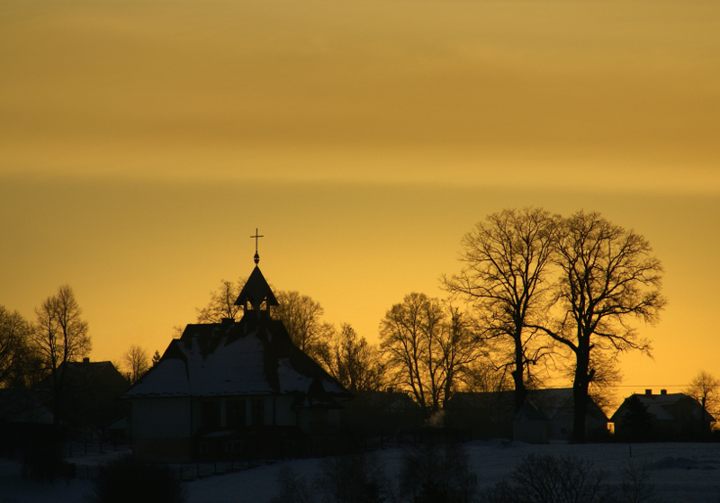 Widok na kościół w Januszowej o zachodzi słońca - niemal czarna sylwetka budynku z dominującym nad nim krzyżem oraz korony drzew pozbawione liści. W tle żółte niebo, dach kościoła pokryty śniegiem
